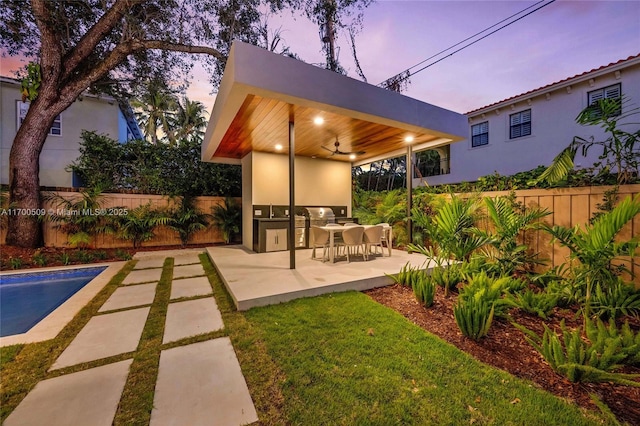 patio terrace at dusk with a fenced in pool, ceiling fan, and exterior kitchen