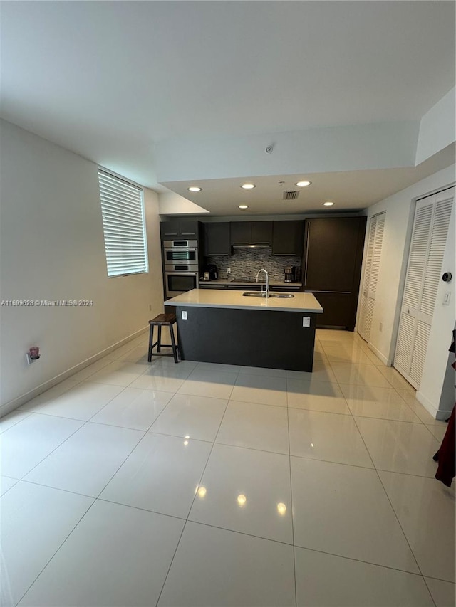 kitchen featuring decorative backsplash, double oven, sink, an island with sink, and light tile patterned flooring