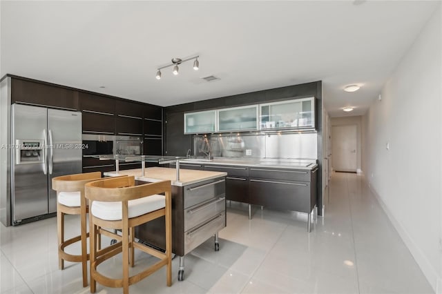 kitchen featuring dark brown cabinetry, tasteful backsplash, stainless steel fridge with ice dispenser, an island with sink, and light tile patterned floors