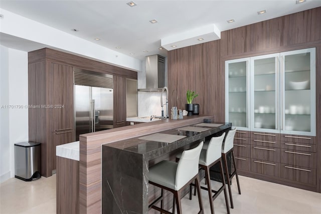 kitchen with wall chimney range hood, sink, built in refrigerator, kitchen peninsula, and a breakfast bar area