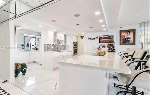 kitchen with decorative backsplash, kitchen peninsula, double oven, white cabinets, and a breakfast bar area