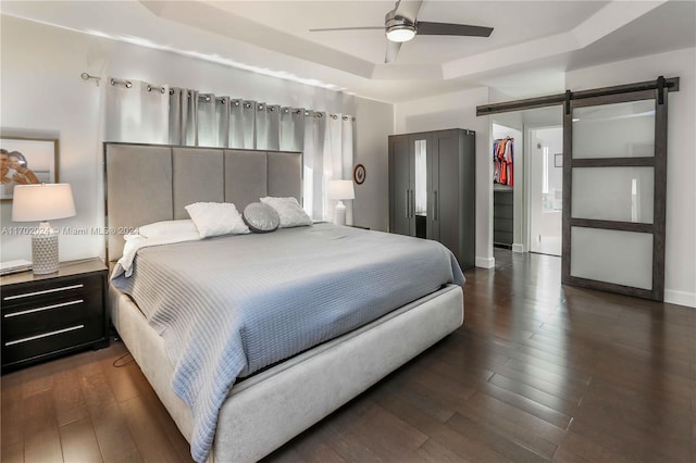 bedroom with dark hardwood / wood-style flooring, a barn door, a tray ceiling, and ceiling fan