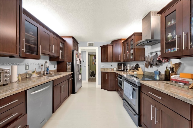 kitchen with sink, wall chimney exhaust hood, light stone countertops, a textured ceiling, and appliances with stainless steel finishes