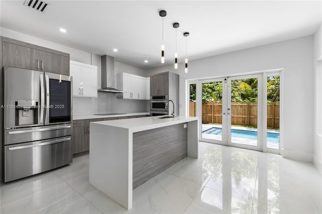 kitchen featuring white cabinetry, stainless steel fridge with ice dispenser, an island with sink, pendant lighting, and wall chimney range hood