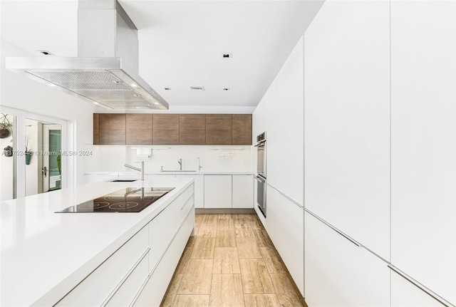 kitchen featuring stainless steel double oven, wall chimney range hood, light hardwood / wood-style flooring, black electric cooktop, and white cabinets