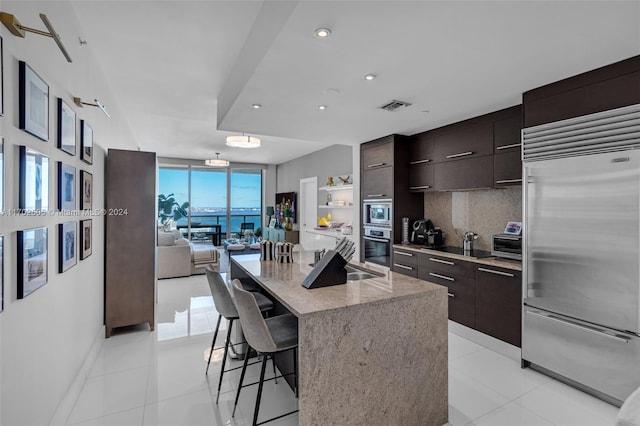 kitchen with a breakfast bar, a kitchen island with sink, built in appliances, decorative backsplash, and dark brown cabinets