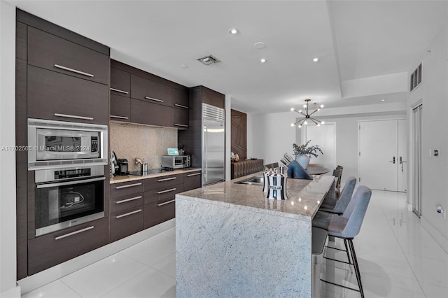 kitchen with a kitchen island with sink, an inviting chandelier, built in appliances, tasteful backsplash, and a breakfast bar area