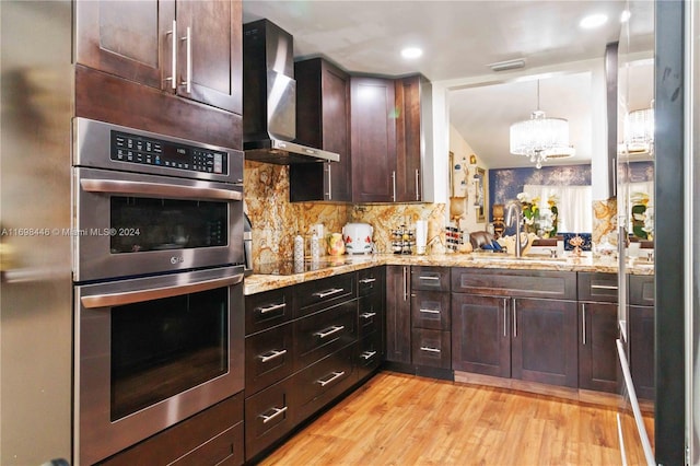 kitchen featuring wall chimney range hood, double oven, a chandelier, black electric cooktop, and light wood-type flooring