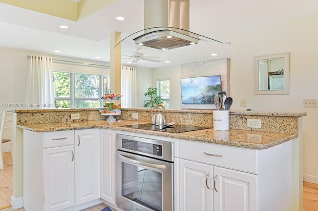 kitchen with stainless steel oven, light hardwood / wood-style flooring, light stone countertops, island range hood, and white cabinetry