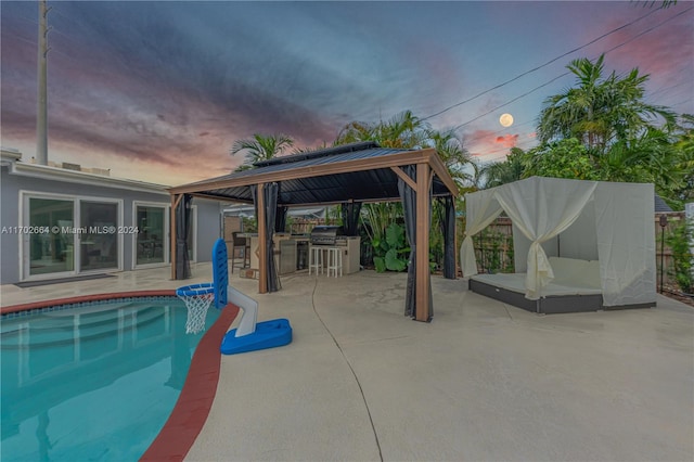 pool at dusk with exterior kitchen, an outdoor bar, a gazebo, a patio area, and area for grilling