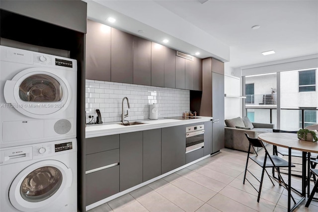 kitchen with gray cabinetry, stainless steel oven, stacked washing maching and dryer, decorative backsplash, and light tile patterned floors