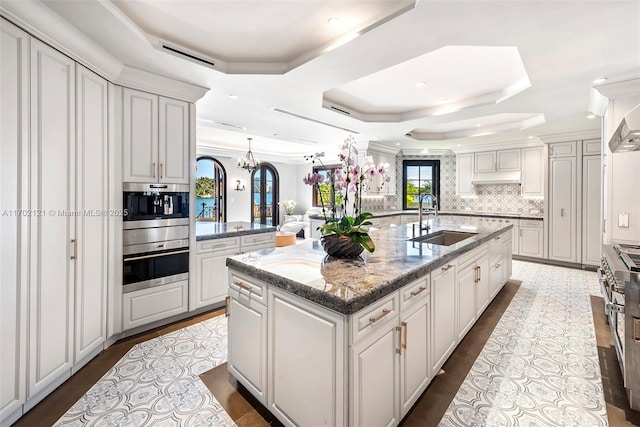 kitchen with white cabinets, an island with sink, sink, backsplash, and a tray ceiling