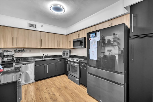 kitchen with light wood-type flooring, stainless steel appliances, sink, light brown cabinets, and dark stone countertops