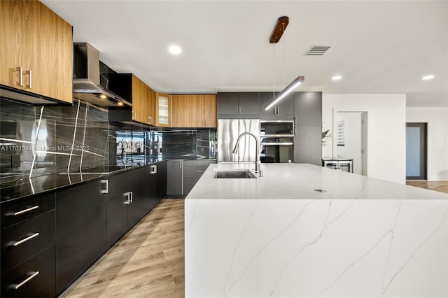 kitchen featuring a center island with sink, stainless steel appliances, a sink, modern cabinets, and wall chimney exhaust hood