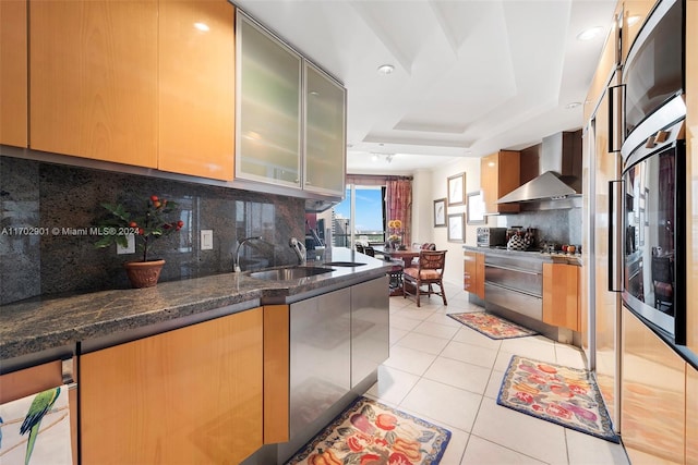 kitchen with sink, wall chimney exhaust hood, a raised ceiling, backsplash, and dark stone counters