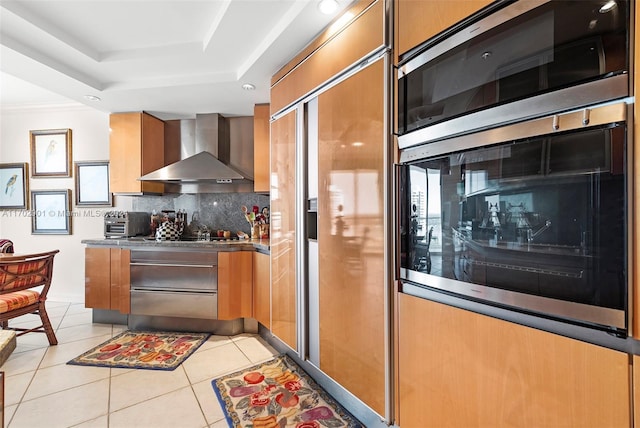 kitchen with backsplash, a raised ceiling, wall chimney range hood, light tile patterned floors, and stainless steel appliances