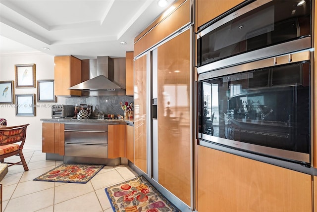kitchen featuring wall chimney exhaust hood, a tray ceiling, brown cabinetry, appliances with stainless steel finishes, and light tile patterned flooring