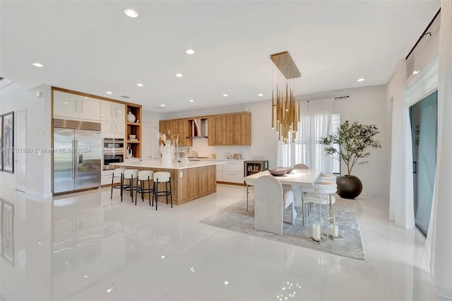 kitchen featuring stainless steel appliances, wall chimney range hood, an island with sink, pendant lighting, and white cabinets