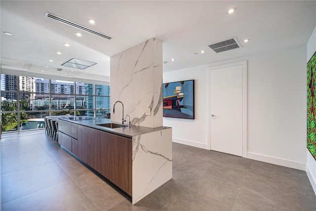 kitchen with light stone countertops, dark brown cabinets, a kitchen island with sink, and sink