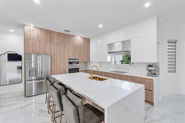 kitchen featuring stainless steel appliances, wall chimney range hood, a kitchen breakfast bar, a kitchen island with sink, and white cabinets