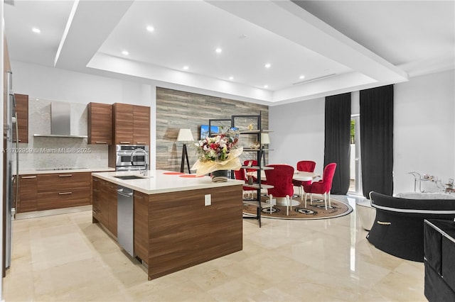 kitchen featuring wall chimney exhaust hood, a center island with sink, a raised ceiling, appliances with stainless steel finishes, and sink