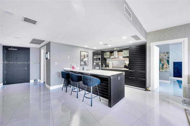 kitchen with a breakfast bar, wall chimney exhaust hood, an island with sink, appliances with stainless steel finishes, and light tile patterned flooring