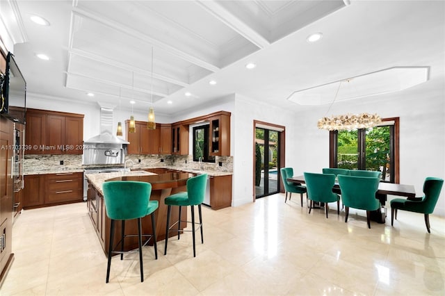 kitchen with coffered ceiling, wall chimney range hood, hanging light fixtures, tasteful backsplash, and beam ceiling