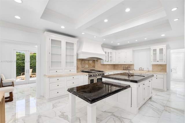 kitchen featuring premium range hood, tasteful backsplash, a center island with sink, range with two ovens, and white cabinetry