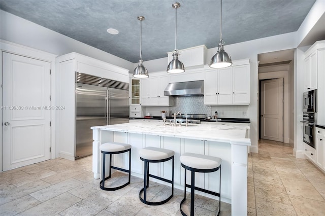 kitchen featuring built in appliances, a kitchen island with sink, decorative light fixtures, and ventilation hood