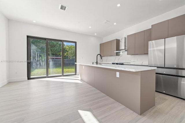 kitchen featuring sink, wall chimney exhaust hood, stainless steel fridge, an island with sink, and light hardwood / wood-style floors