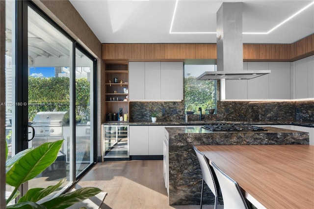 kitchen featuring island exhaust hood, light wood-type flooring, stainless steel gas cooktop, white cabinets, and wine cooler