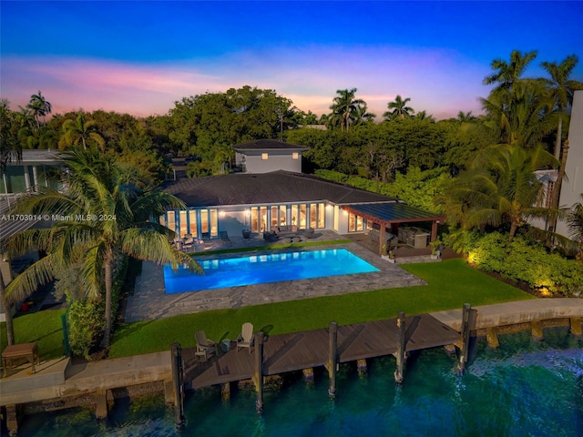 pool at dusk with a patio, a water view, and a lawn