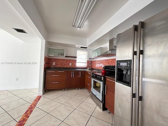 kitchen featuring light tile patterned flooring, wall chimney range hood, backsplash, and appliances with stainless steel finishes