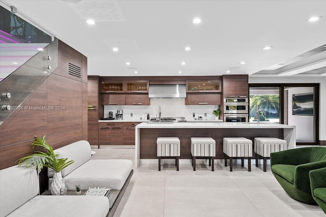 kitchen featuring a breakfast bar area, a kitchen island with sink, dark brown cabinets, and wall chimney exhaust hood