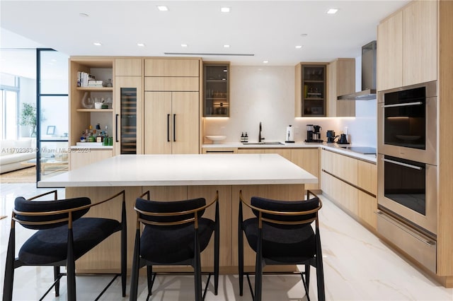 kitchen featuring a center island, light brown cabinets, wall chimney exhaust hood, sink, and a kitchen bar