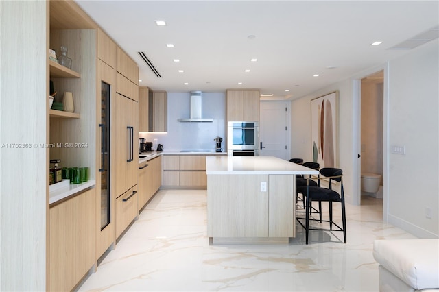 kitchen with a breakfast bar, a center island, wall chimney exhaust hood, double oven, and light brown cabinetry