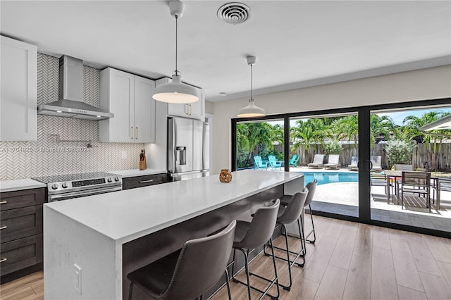 kitchen with light wood-type flooring, wall chimney exhaust hood, stainless steel appliances, decorative light fixtures, and white cabinetry