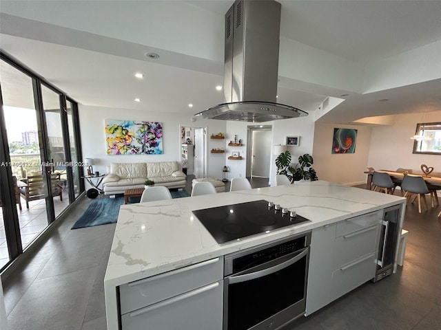 kitchen with black electric stovetop, oven, a kitchen island, island range hood, and light stone counters