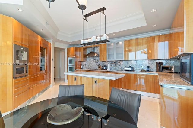 kitchen featuring a raised ceiling, backsplash, decorative light fixtures, and wall chimney range hood