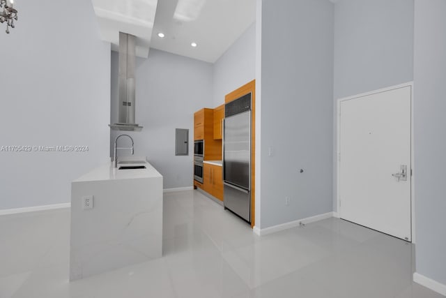 kitchen with wall chimney range hood, sink, built in refrigerator, light tile patterned floors, and a towering ceiling