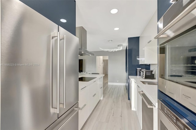 kitchen with appliances with stainless steel finishes, sink, wall chimney range hood, light hardwood / wood-style flooring, and white cabinets