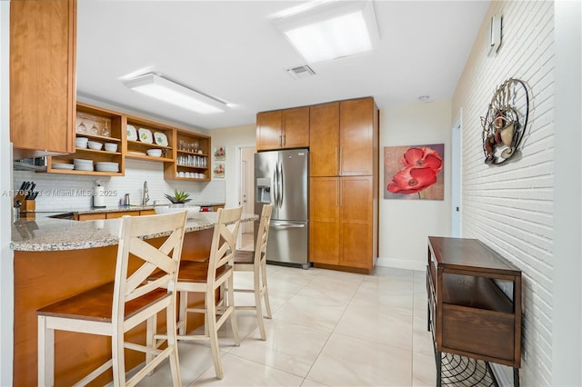 kitchen featuring open shelves, brown cabinetry, stainless steel fridge with ice dispenser, decorative backsplash, and light stone countertops