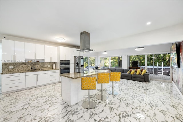 kitchen featuring a kitchen bar, dark stone countertops, appliances with stainless steel finishes, island exhaust hood, and white cabinets