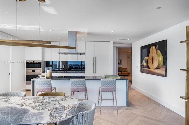 kitchen with stainless steel double oven, island range hood, sink, white cabinets, and light parquet flooring