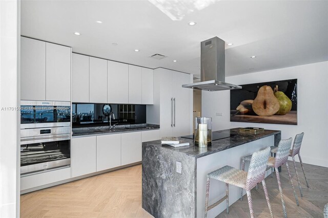 kitchen with dark stone counters, light parquet floors, double oven, wall chimney range hood, and white cabinets