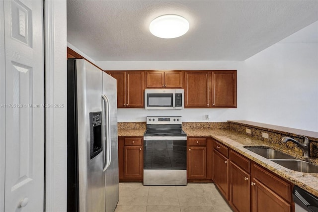 kitchen with a textured ceiling, light stone counters, stainless steel appliances, a sink, and brown cabinets