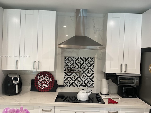 kitchen with wall chimney range hood, light stone counters, backsplash, black electric cooktop, and white cabinets