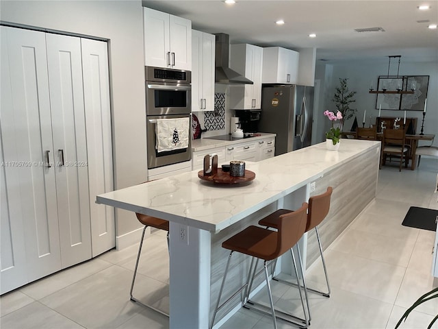 kitchen featuring white cabinetry, a center island, wall chimney exhaust hood, stainless steel appliances, and a kitchen bar