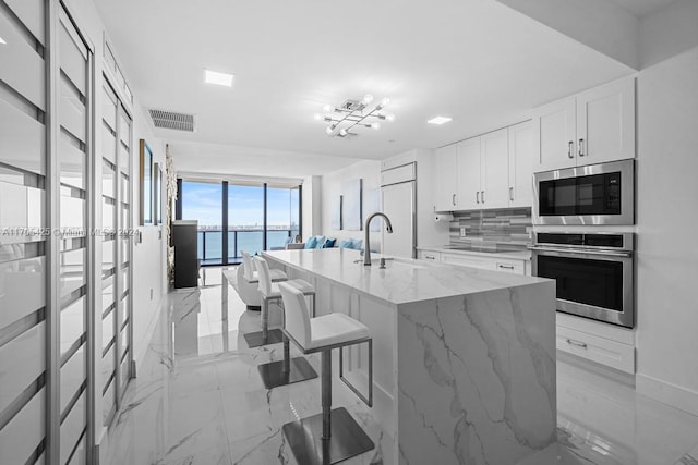 kitchen with a center island with sink, a water view, built in appliances, light stone countertops, and a kitchen bar