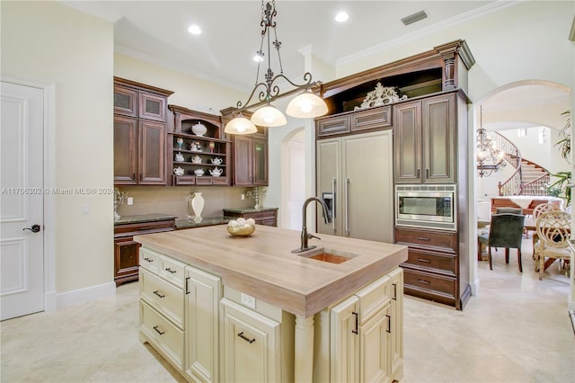 kitchen with a kitchen island with sink, cream cabinets, wooden counters, sink, and built in appliances